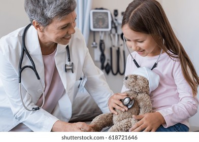 Happy Little Girl Checking Heartbeat Of Teddy Bear In Hospital. Happy Pediatrician And Child Patient Playing. Girl And Senior Doctor With Stethoscope Auscultate The Heartbeat Of The Ill Teddy Bear.