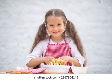 Happy little girl in an  chef costume helps to cook a pie by kneading the dough, a child smiles, cooking according to a recipe, a girl helps to cook for mom, a child's portrait and development.
