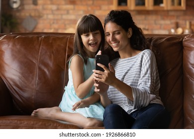 Happy Little Girl Child Relax On Sofa At Home With Young Latino Mother Talk On Video Call On Cellphone. Smiling Hispanic Mom With Small Daughter Have Fun Engaged In Webcam Online Digital Conversation.