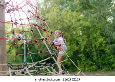 A happy little girl is climbing on an outdoor playground on a summer day. Blonde girl climbs to the top of the game complex - Powered by Shutterstock