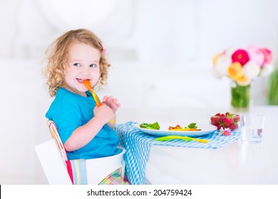 Happy Little Girl, Cute Curly Toddler, Eating Fresh Vegetables For Lunch, Healthy Salad Snack, Corn, Broccoli, Carrots And Strawberry Fruit In A White Dining Room Sitting In A High Chair