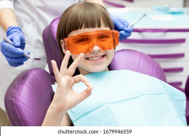 Happy little girl at dentist appointment. Kid gesturing okay sign with hand. Child wearing orange protective plastic glasses sit on dental chair. Qualified pediatric stomatological service approval