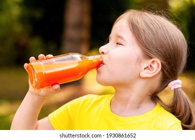 Happy little girl drinking healthy orange carrot juice from a glass bottle delighted, portrait, face closeup, outdoors. Healthy drink, nutritious vegetable juice, drink full of vitamins lifestyle shot