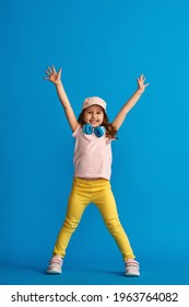 happy little girl in fashionable summer clothes and headphones stands on blue background in full growth. child poses with his hands raised in new concept summer and spring casual children's clothing.