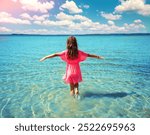 Happy little girl with hands in the air walks on the beach in summer back to camera in daylight. The girl in a red dress looks at the sea on a sunny day.