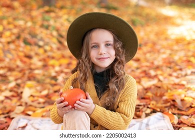 Happy Little Girl In Hat And Autumn Clothes On An Autumn Background. A Smiling Child Is Playing In Fall Park. Young Kid Girl Has Fun Playing With Falling Golden Leaves. Autumn Portrait Of Cute Girl
