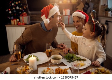 Happy little girl having fun and feeding her father while having Christmas lunch at dining table. 