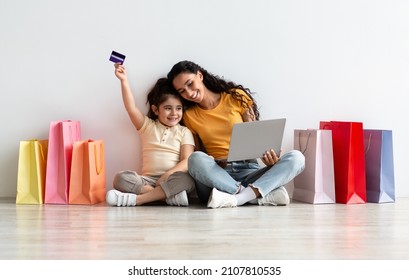 Happy Little Girl And Her Mom Making Online Shopping With Laptop And Credit Card At Home, Excited Mother And Female Child Sitting On Floor Among Bright Shopper Bags, Enjoying Internet Purchases