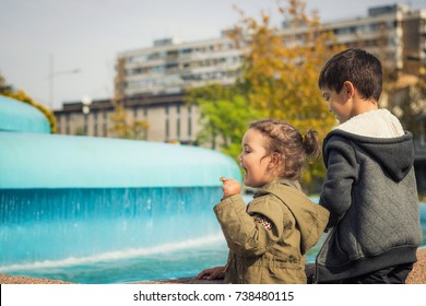 Happy Little Girl Holding Coin And Making A Wish While Standing By The Fountain With Her Brother. Children Throwing Coins Into Fountain.