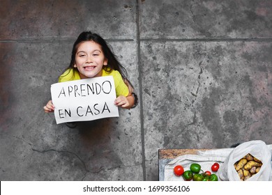 Happy little girl holding a sheet of paper that says in spanish "STUDYING AT HOME".