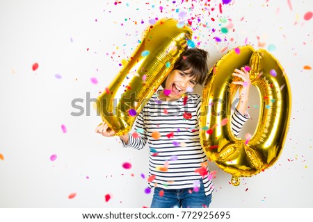 Happy little girl holding two golden balloons making the 10 number while falling confetti on white background. 10th anniversary celebration party.