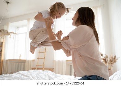 Happy Little Girl Jumping In Bed Holds Mom Hands View From Bottom, Fit Family Funny Leisure Activity At Home Daughter And Mother Priceless Time Together In Morning After Night Or Daytime Sleep Concept