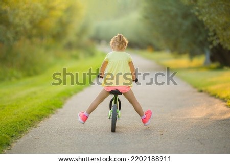 Happy little girl lifting legs up and riding on first bike without pedals on sidewalk at city park in warm summer day. 3 years old toddler. Back view. Learning to keep balance.