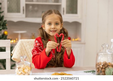 Happy little girl with a lollipop near Christmas tree at home. Little girl with sweet candies on Xmas eve. Family, tradition, celebration concept. Candy canes in shape of heart in hand child - Powered by Shutterstock