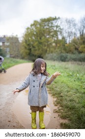 Happy Little Girl Looking Adorably At Her Muddy Clothes After Jumping In Puddle. Young Child Having Fun And Getting Dirty In Puddles. Kids Playing Outside