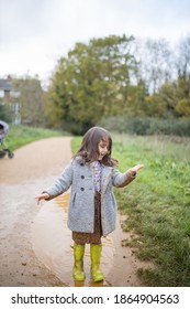 Happy Little Girl Looking Adorably At Her Muddy Clothes After Jumping In Puddle. Young Child Having Fun And Getting Dirty In Puddles. Kids Playing Outside