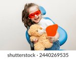 Happy little girl looking in the mirror at her teeth after teeth surgery in a pediatric stomatology. A little girl sits in a dental chair and examines her cured teeth while looking in the mirror