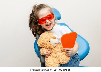 Happy little girl looking in the mirror at her teeth after teeth surgery in a pediatric stomatology. A little girl sits in a dental chair and examines her cured teeth while looking in the mirror - Powered by Shutterstock