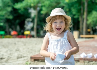 Happy Little Girl Making Paper Boat On The Beach. Vacation Concept. Paper Ship In Child Hands. Preschool Child In White T-shirt And Hat On Sunny Day. 