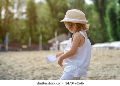 Happy Little Girl Making Paper Boat On The Beach. Vacation Concept. Paper Ship In Child Hands. Preschool Child In White T-shirt And Hat On Sunny Day. 