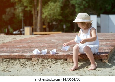 Happy Little Girl Making Paper Boat On The Beach. Vacation Concept. Paper Ship In Child Hands. Preschool Child In White T-shirt And Hat On Sunny Day. 