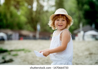 Happy Little Girl Making Paper Boat On The Beach. Vacation Concept. Paper Ship In Child Hands. Preschool Child In White T-shirt And Hat On Sunny Day. 