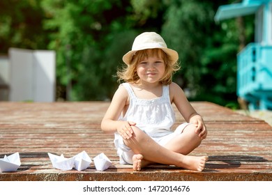 Happy Little Girl Making Paper Boat On The Beach. Vacation Concept. Paper Ship In Child Hands. Preschool Child In White T-shirt And Hat On Sunny Day. 