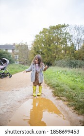 Happy Little Girl With Muddy Clothes Smiling And Looking Down At Puddle Before Jumping In It. Young Child Having Fun And Getting Dirty In Puddles. Kids Playing Outside