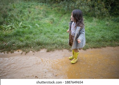 Happy Little Girl With Muddy Clothes Smiling And Looking At Puddle Before Jumping In It. Young Child Having Fun Jumping In Puddles. Kids Playing Outside