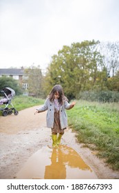 Happy Little Girl With Muddy Clothes Smiling And Looking At Puddle Before Jumping In It. Young Child Having Fun Jumping In Puddles. Kids Playing Outside