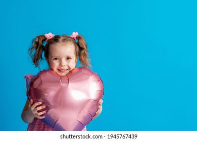 happy little girl in pink dress and two ponytails for her birthday is holding pink balloon, in shape heart, isolated on blue background. Advertising, holiday. Positive children's emotions. Copy space