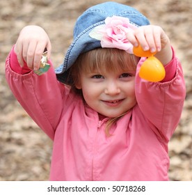 A Happy Little Girl In Pink With A Hat Opens An Orange Easter Egg To Get The Candy Inside During Egg Hunt.