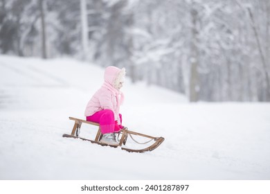 Happy little girl in pink warm clothes sitting on sledge and sledding down on snow from hill. Child enjoying white winter day at park. Side view. - Powered by Shutterstock