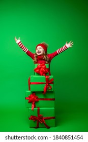 happy little girl in red hat plays with pyramid Christmas gift boxes on green background in Studio. child is happy to close his eyes and raise his hands up, gets lot gifts for Christmas. Advertising.