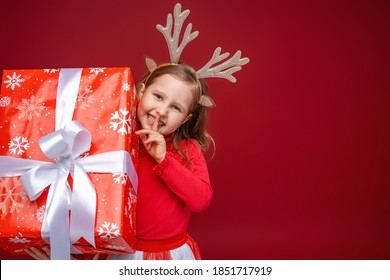 Happy little girl in reindeer antlers on a red background. a cheerful child, peeking out from behind box and pressing finger to his mouth. Secret. Christmas sales, advertising, discounts. Copy space.