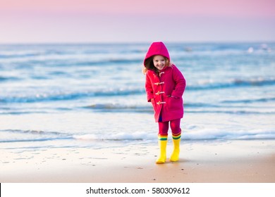 Happy Little Girl Running And Jumping In The Waves On North Sea Beach During Winter Vacation In Holland. Kids Play In Ocean Sand Dunes On Cold Autumn Or Spring Day. Beach Fun For Family With Children