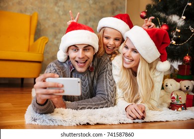 Happy little girl in Santa's hat makes horns to her parents while her father taking Christmas photo on mobile phone Stock Photo