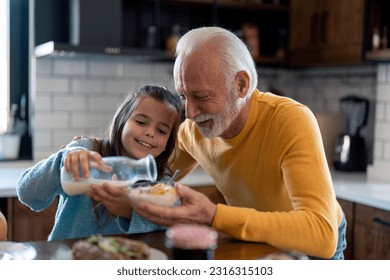 Happy little girl with senior grandfather preparing morning meal in the kitchen at home, adding milk into bowl with cereals, enjoying their time. - Powered by Shutterstock