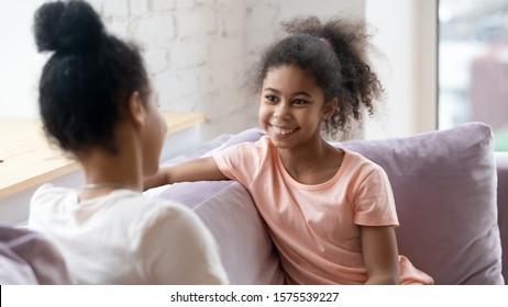 Happy Little Girl Sit On Couch With Young Mom Or Nanny Talking Or Chatting, Gossiping On Weekend At Home, Smiling Small Child Relax On Sofa With Mother, Share Secrets With Parent, Bonding Concept