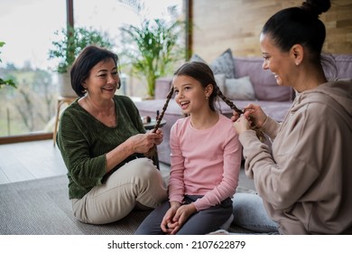 Happy little girl sitting on floor, her mother and grandmother making her plaits at home.