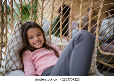 Happy little girl sitting in wicker rattan hang chair outdoors in patio in autumn, looking at camera