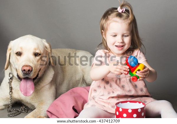 Happy\
little girl with smiling funny face playing with plastic car toy\
near labrador dog pet in studio on grey\
background