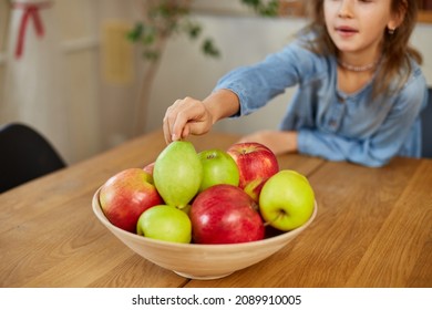 Happy little girl takes a pear from the bowl with variety fruits on table at home, child eating healthy snack, vegetarian nutrition for kids, vitamins for children.