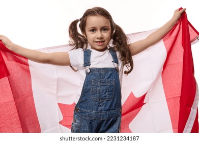 Happy little girl with two ponytails, wearing blue denim overalls, carries Canadian flag. Concept of Independence Day of Canada, the first July. Immigration. Emigration. Travel and tourism concept - Shutterstock ID 2231234187