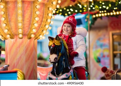 Happy Little Girl In Warm Jacket And Red Knitted Nordic Hat And Scarf Riding Carousel Horse During Family Trip To Traditional German Christmas Market. Kids At Xmas Outdoor Fair On Snowy Winter Day.