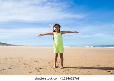 Happy little girl wearing summer cloth, standing with open arms on golden sand, looking at camera, spending leisure time on beach at sea. Full length, front view. Childhood concept
