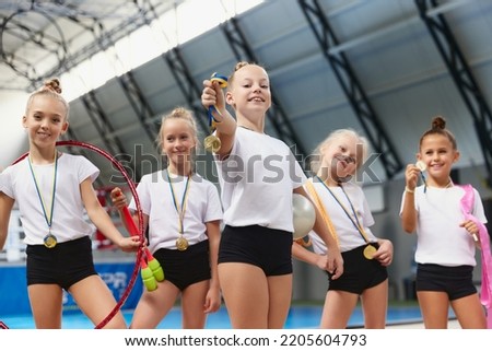 Happy little girls, female gymnastics athletes showing their medals at camera at sports gym, indoors. Concept of win, sport, studying, achievements and success. Teamwork, friendship and support