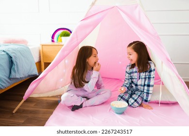Happy little girls in pajamas playing during a fun sleepover eating popcorn while talking in a pink teepee