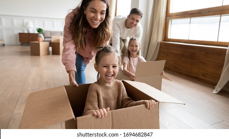 Happy little girls sisters sitting in big carton boxes while joyful parents pushing it on floor. Laughing family couple having fun with small children in new house, relocation moving day concept.