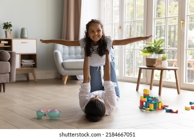 Happy Little Indian Daughter Girl And Strong Mom Doing Acroyoga At Home, Playing Airplane Flight, Smiling, Laughing. Mother Holding Child In Arms, Lifting Girl Up In Air, Doing Active Exercises